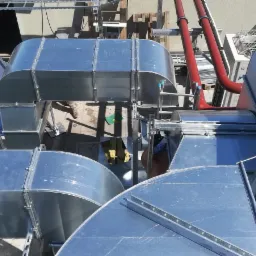 Widok z góry na system kanałów wentylacyjnych z blachy ocynkowanej, z widocznymi elementami montażowymi i rurami w kolorze czerwonym na dachu budynku.