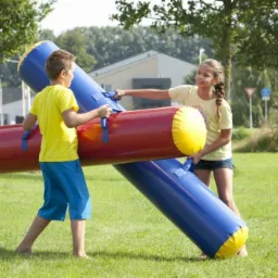 Dwoje dzieci, chłopiec i dziewczynka, bawi się na trawie z dużymi, kolorowymi dmuchanymi walcami połączonymi ze sobą.