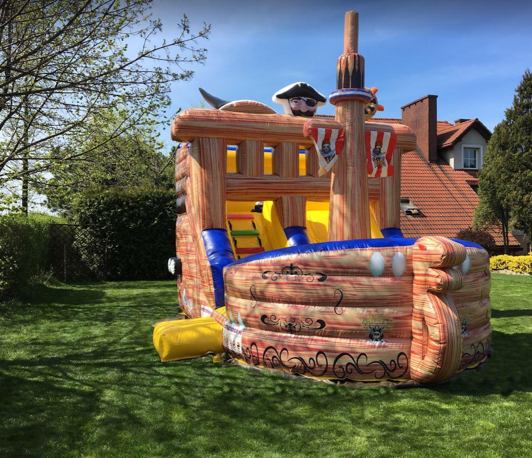 Nadmuchiwania atrakcja dla dzieci w kształcie pirackiego statku z drabinką i zjeżdżalnią, ustawiona na trawniku przed domem z czerwoną dachówką.