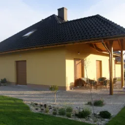 Żółty dom jednorodzinny z czarnym dachem z dachówki, drewnianą pergolą nad tarasem, oraz starannie zagospodarowanym ogrodem z trawnikiem i kamienną ścieżką.