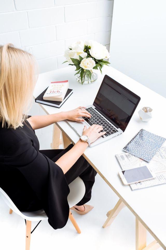 Kobieta w czarnym stroju pisze na laptopie przy białym biurku z wazonem białych kwiatów, książką 'Think and Grow Rich', notatnikiem i smartfonem.