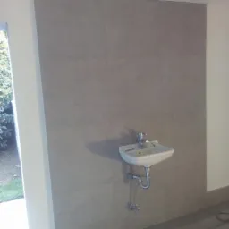 Umywalka zamontowana na ścianie wyłożonej szarymi płytkami w łazience, widoczny fragment podłogi i okno z widokiem na zieleń.