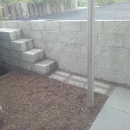 Wykonana z jasnoszarej kostki brukowej ściana oporowa z wbudowanymi schodami, prowadzącymi do wyżej położonego terenu, obok trawnika i słupka oświetleniowego.