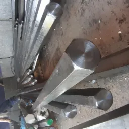 Wiązka sześciokątnych prętów stalowych ułożonych w magazynie, widoczne ślady korozji i zabrudzenia na powierzchni.