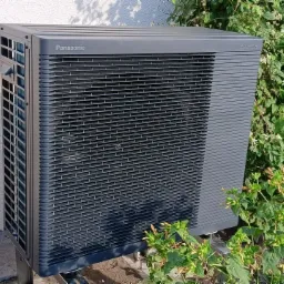 Szara pompa ciepła Panasonic Aquarea zainstalowana przy ścianie budynku w Sępólnie Krajeńskim, otoczona zielenią. Widok z boku, oświetlenie dzienne.