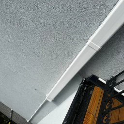 Ekover- Piotr Drabjewski - Biała rura PCV przymocowana do szarej ściany budynku, widoczna drabina i fragment balkonu z drewnianą podłogą i metalową balustradą. Ujęcie z dołu.