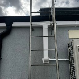 Ekover- Piotr Drabjewski - Metalowa drabina przymocowana do ściany budynku, obok rynna i klimatyzator. Widoczne rury instalacyjne. Pochmurne niebo w tle.