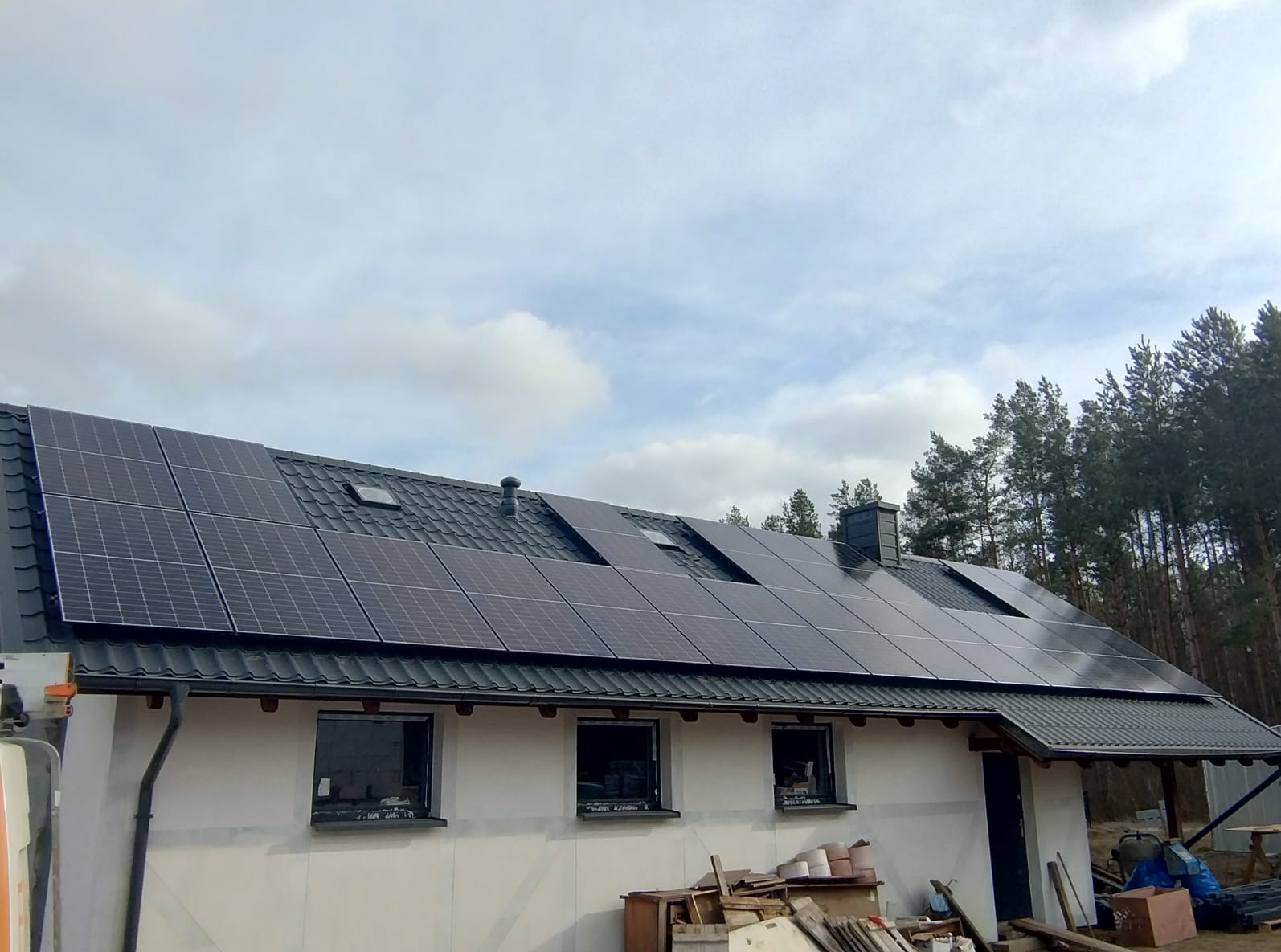 Dom z panelami fotowoltaicznymi na dachu z dachówki, widok z dołu, fragment lasu w tle. Panele ułożone rzędami, widać okna dachowe i komin.