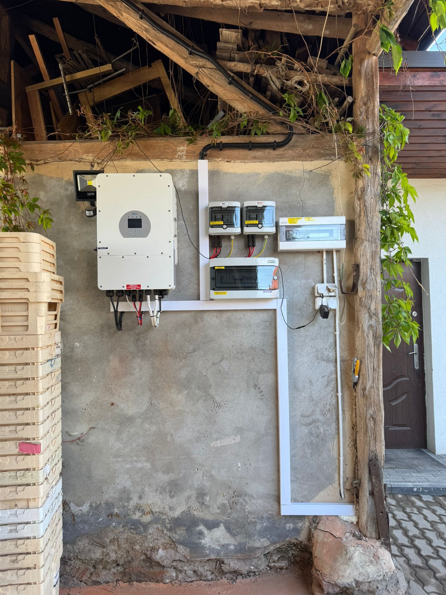 Zamontowany inwerter fotowoltaiczny z rozdzielnią na szarej, chropowatej ścianie. Widoczne skrzynki, kable i elementy instalacji. Drewniana konstrukcja dachu i pnącza.