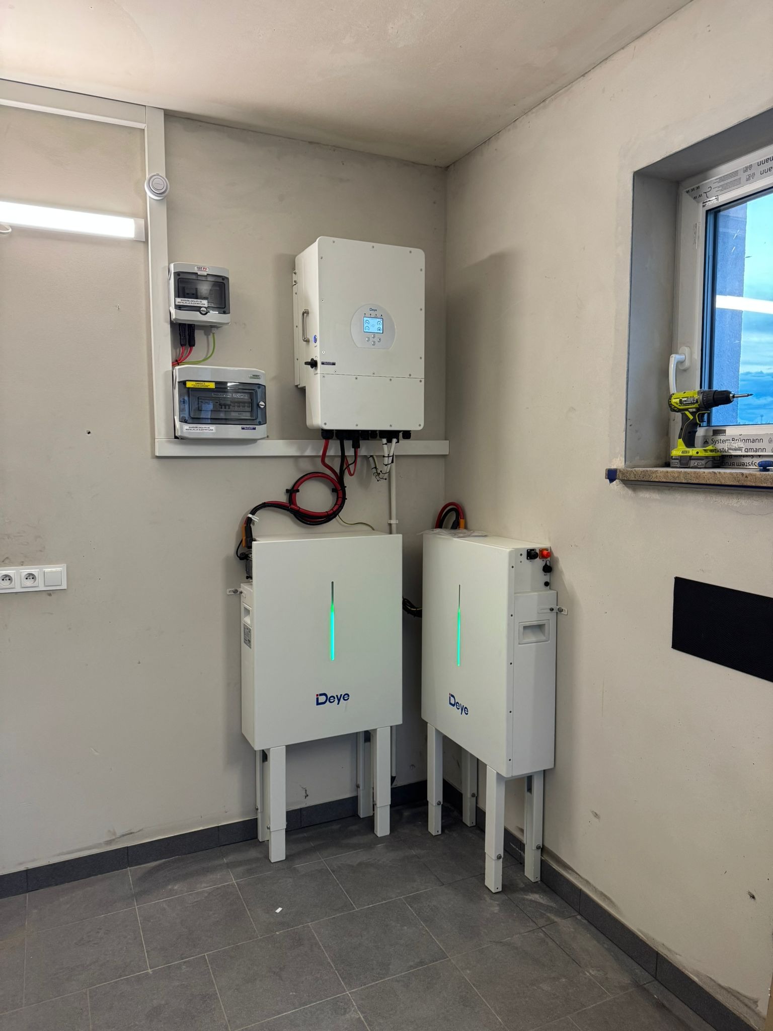 Wnętrze z zamontowanymi dwoma białymi magazynami energii Deye z zielonymi wskaźnikami, inwerterem i rozdzielnicą, w pobliżu okna z wkrętarką.