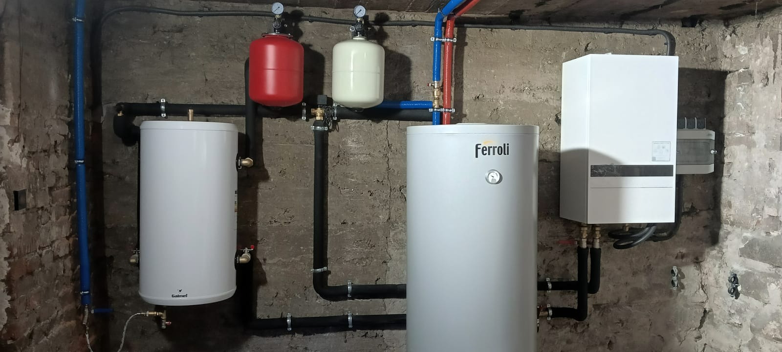 Kompletna instalacja grzewcza: dwa białe zbiorniki, czerwony i biały zbiornik wyrównawczy, piec Ferroli, rury w piwnicy z surowymi ścianami.