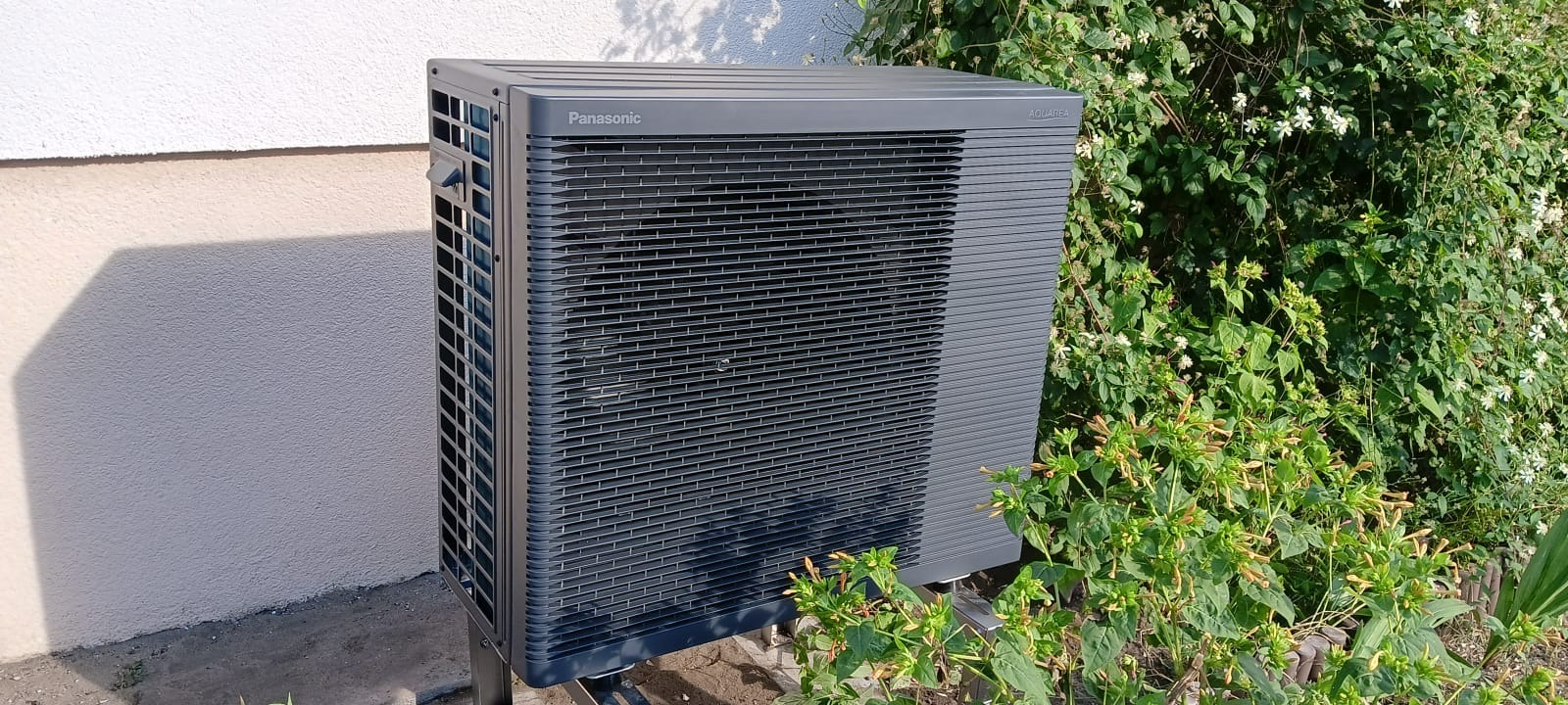 Szara pompa ciepła Panasonic Aquarea zainstalowana przy ścianie budynku w Sępólnie Krajeńskim, otoczona zielenią. Widok z boku, oświetlenie dzienne.