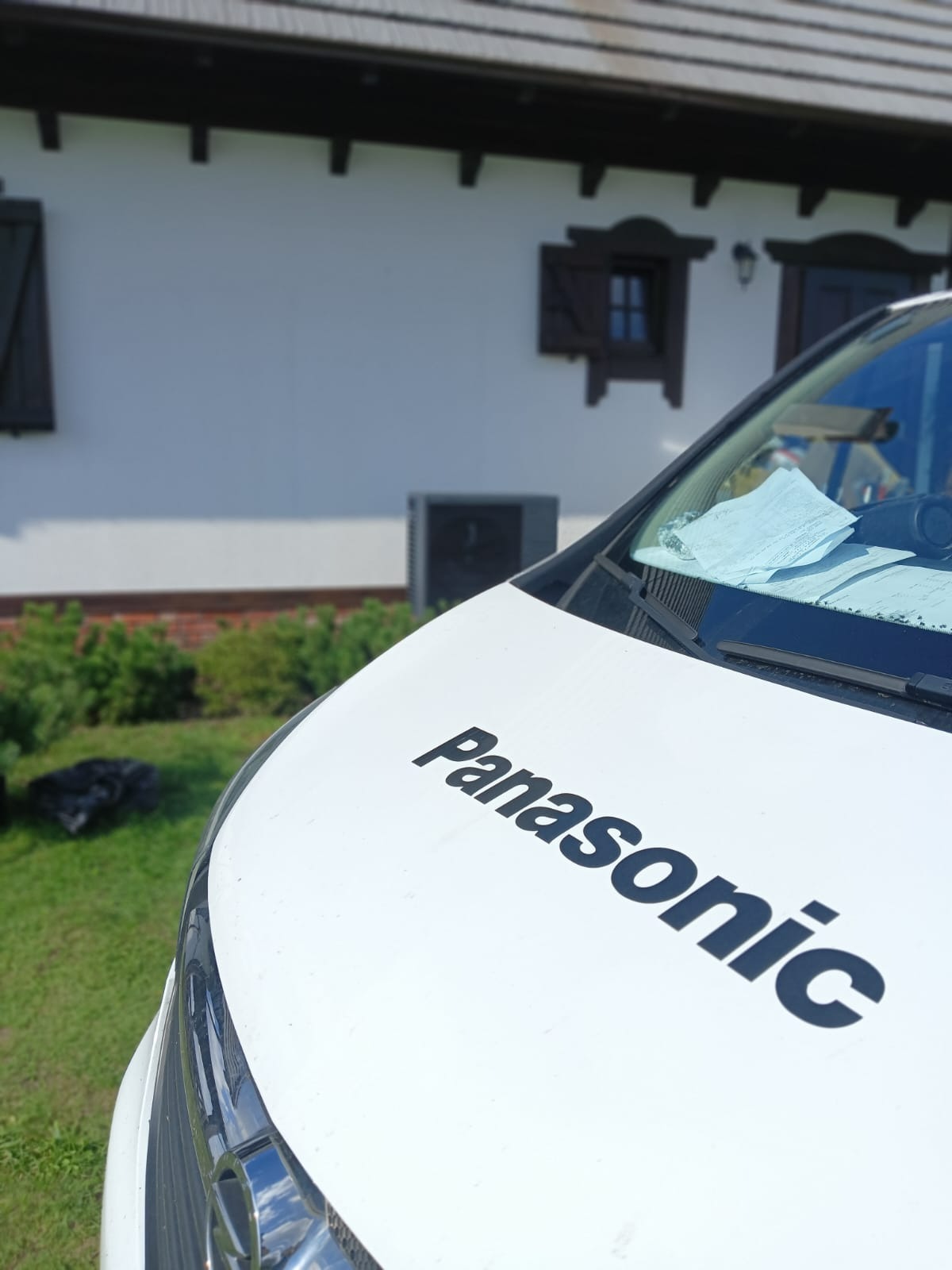 Furgonetka Panasonic na tle domu z pompą ciepła. Widoczne dokumenty na desce rozdzielczej. Usługi instalacji i serwisu OZE.