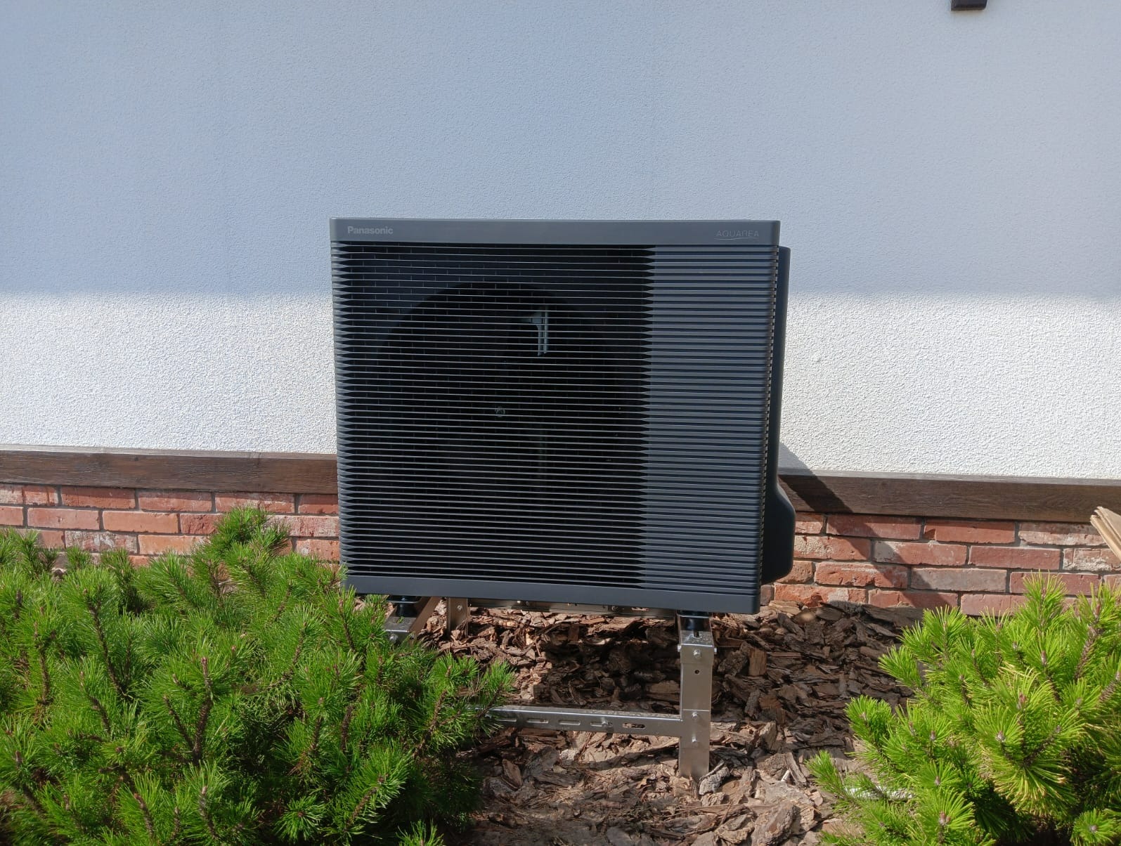 Zewnętrzna jednostka pompy ciepła Panasonic Aquarea na metalowej konstrukcji montażowej, z widocznym logo, na tle białej ściany i zielonych krzewów.