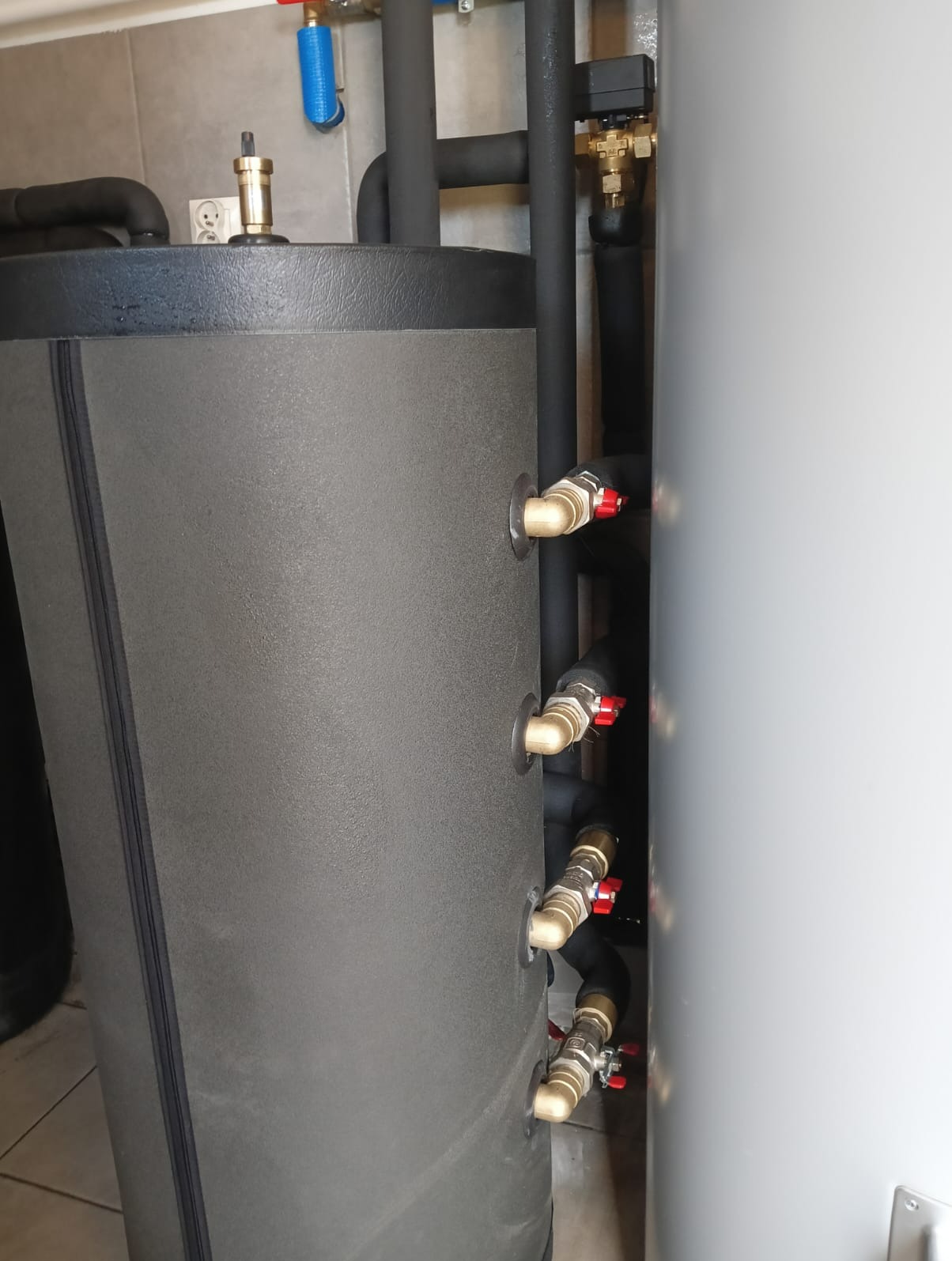Szary zbiornik akumulacyjny z czarnymi rurami i zaworami z czerwonymi pokrętłami, widoczne przyłącza. Element instalacji grzewczej w pomieszczeniu.