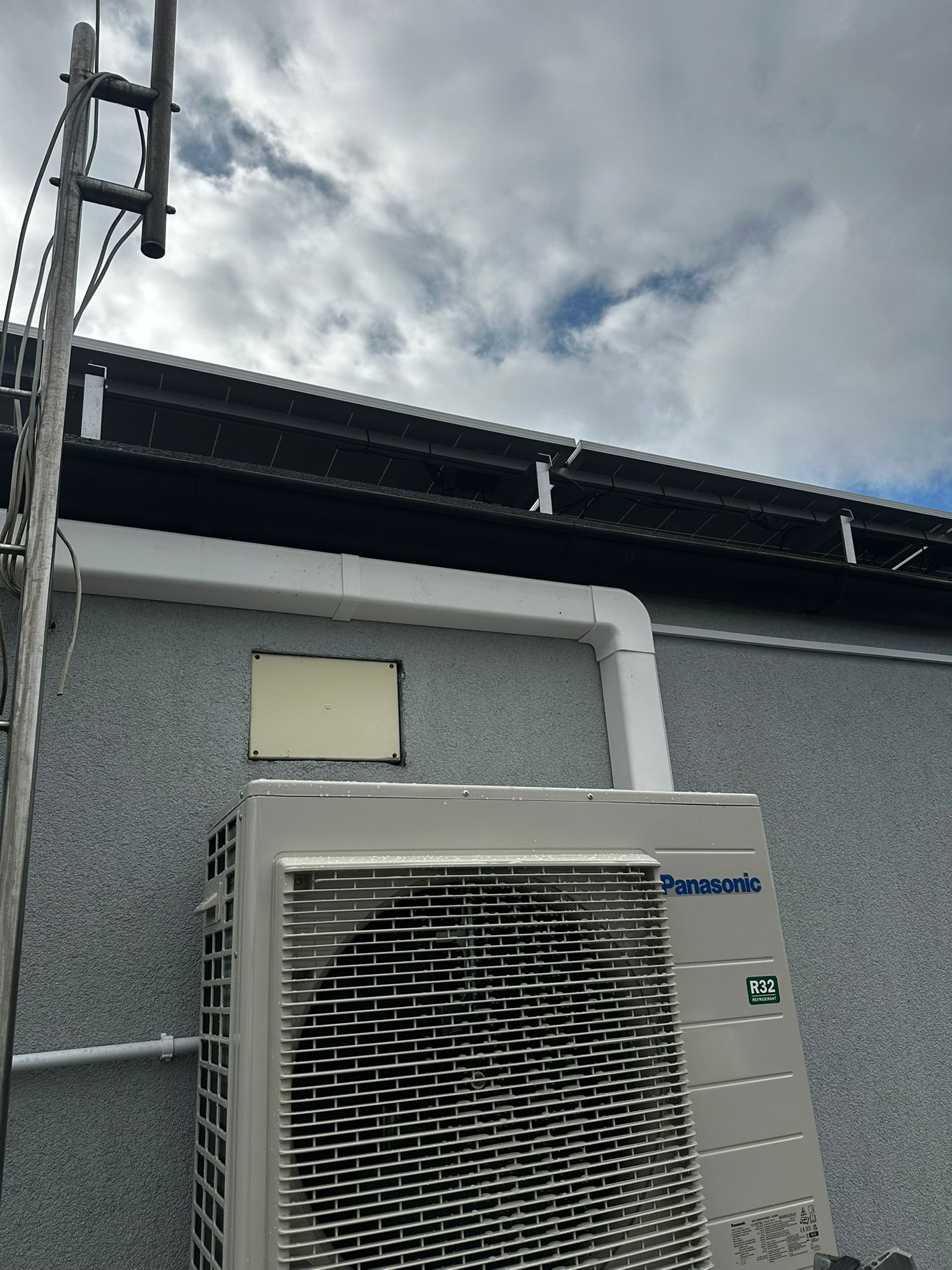 Zewnętrzna jednostka pompy ciepła Panasonic R32 na szarej elewacji budynku, widoczna drabina i fragment dachu z panelami, pochmurne niebo w tle.