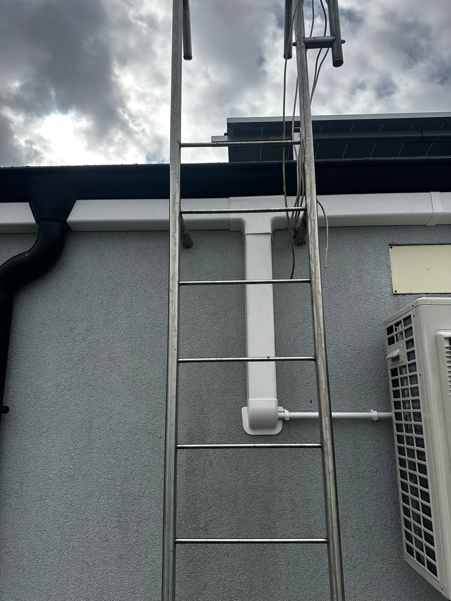 Metalowa drabina przymocowana do ściany budynku, obok rynna i klimatyzator. Widoczne rury instalacyjne. Pochmurne niebo w tle.