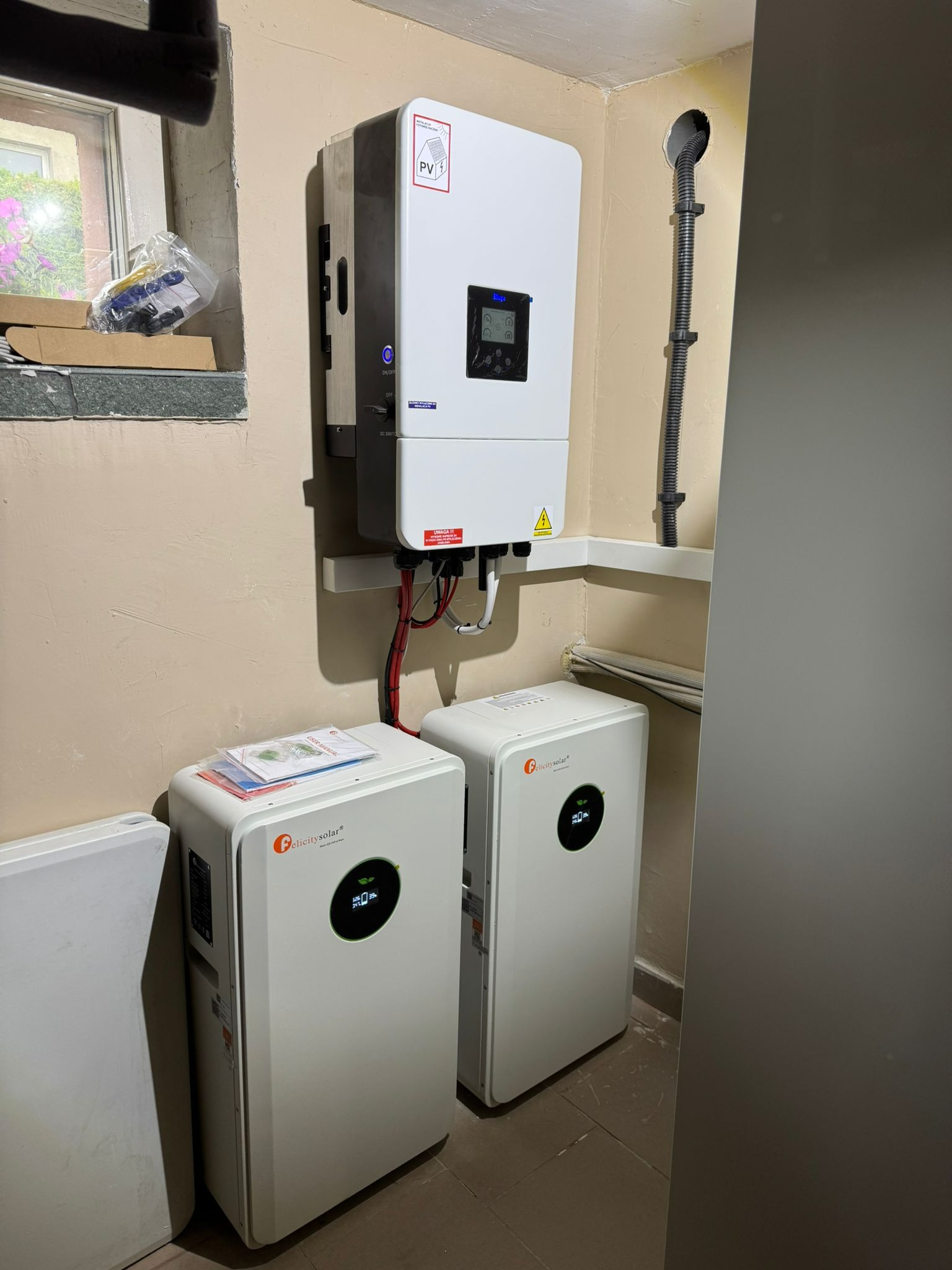 Wnętrze z zamontowanym inwerterem i dwoma akumulatorami energii Felicity Solar na beżowej ścianie. Widoczne okno i przewody. Instalacja w pomieszczeniu.