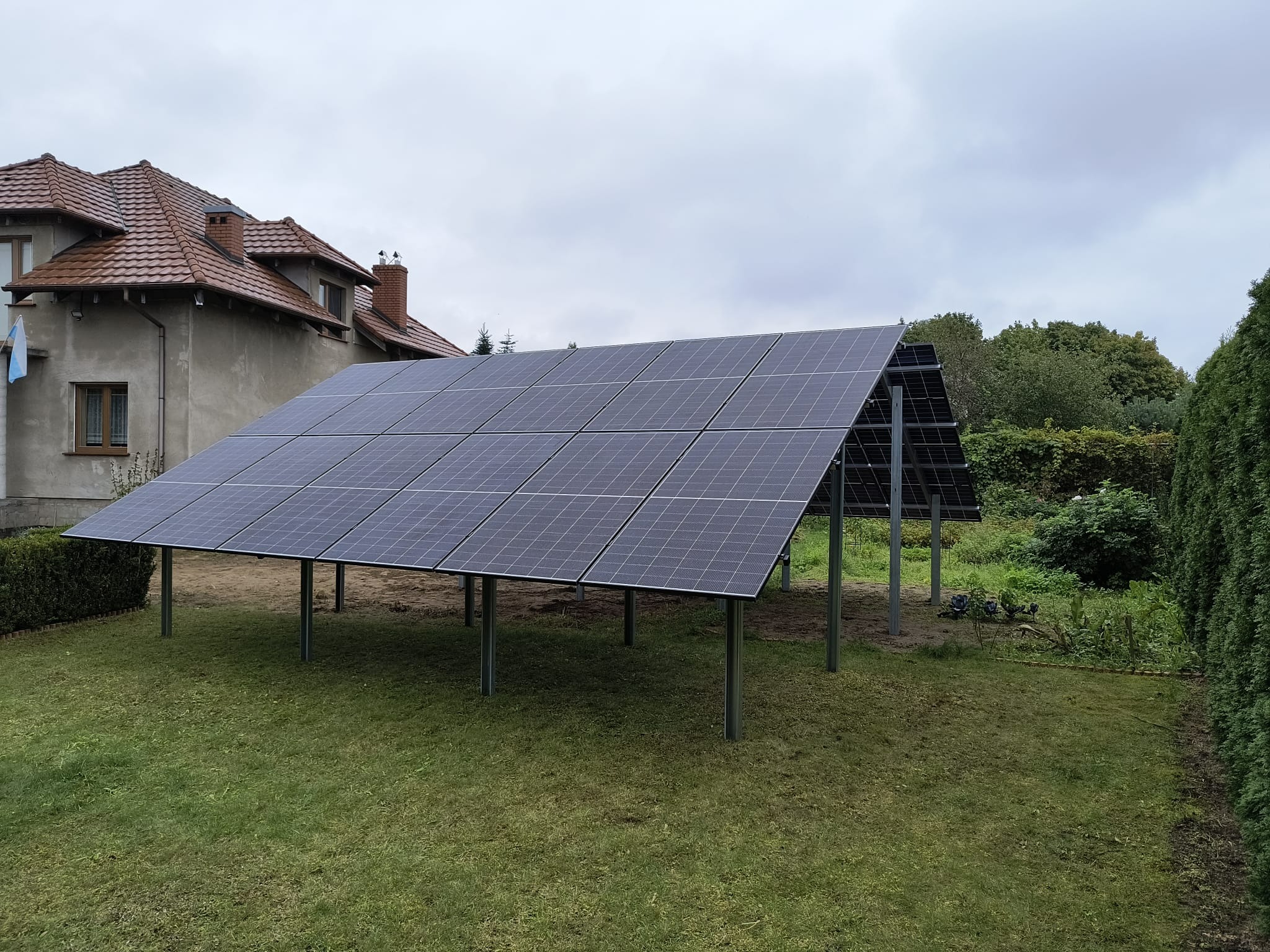 Instalacja fotowoltaiczna na trawniku z metalową konstrukcją wsporczą. W tle dom z czerwoną dachówką i zielona roślinność. Pochmurne niebo.