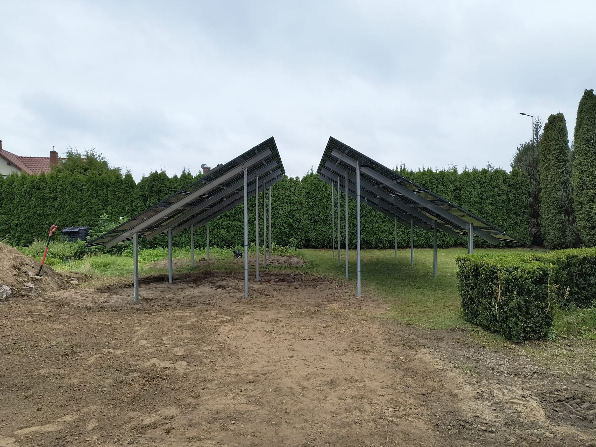 Montaż konstrukcji pod panele fotowoltaiczne na działce z zielenią. Widoczne metalowe słupki i częściowo zamontowane panele. Prace w toku, przygotowanie do instalacji.