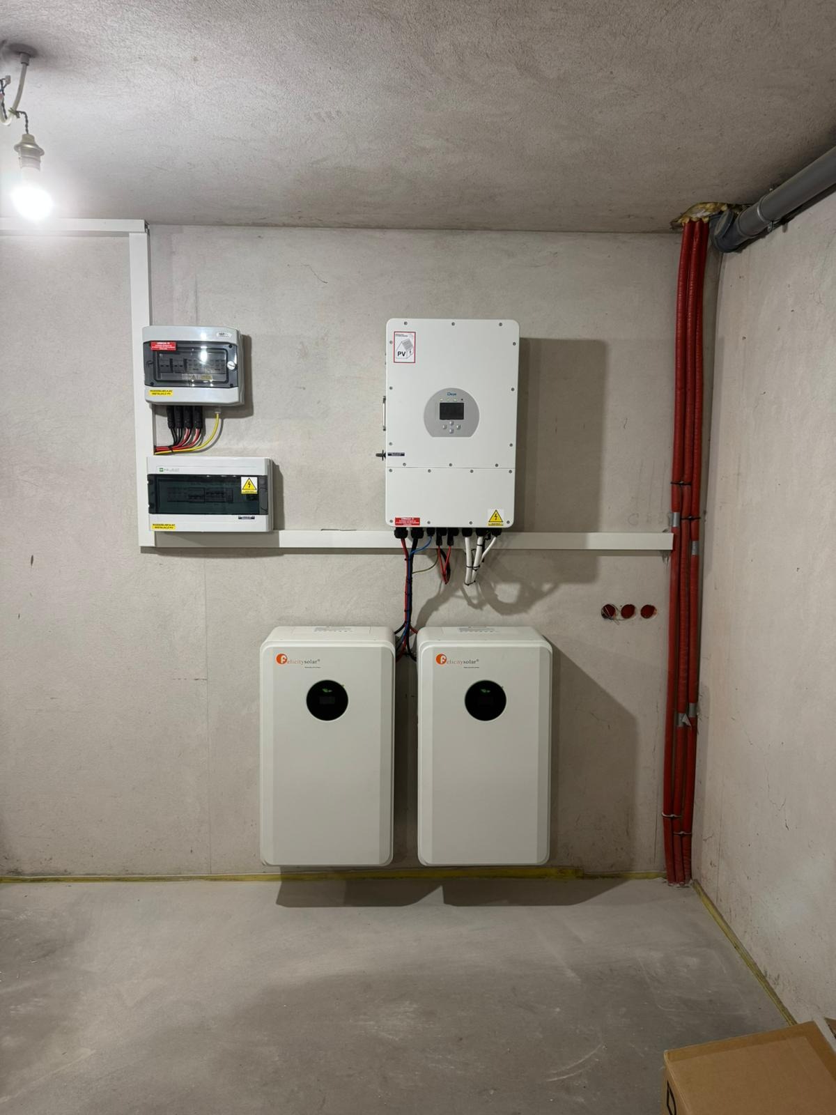 Kompletna instalacja fotowoltaiczna w piwnicy: dwa białe magazyny energii Felicitysolar, inwerter i rozdzielnice na szarej, surowej ścianie. Czerwone kable w peszlu.