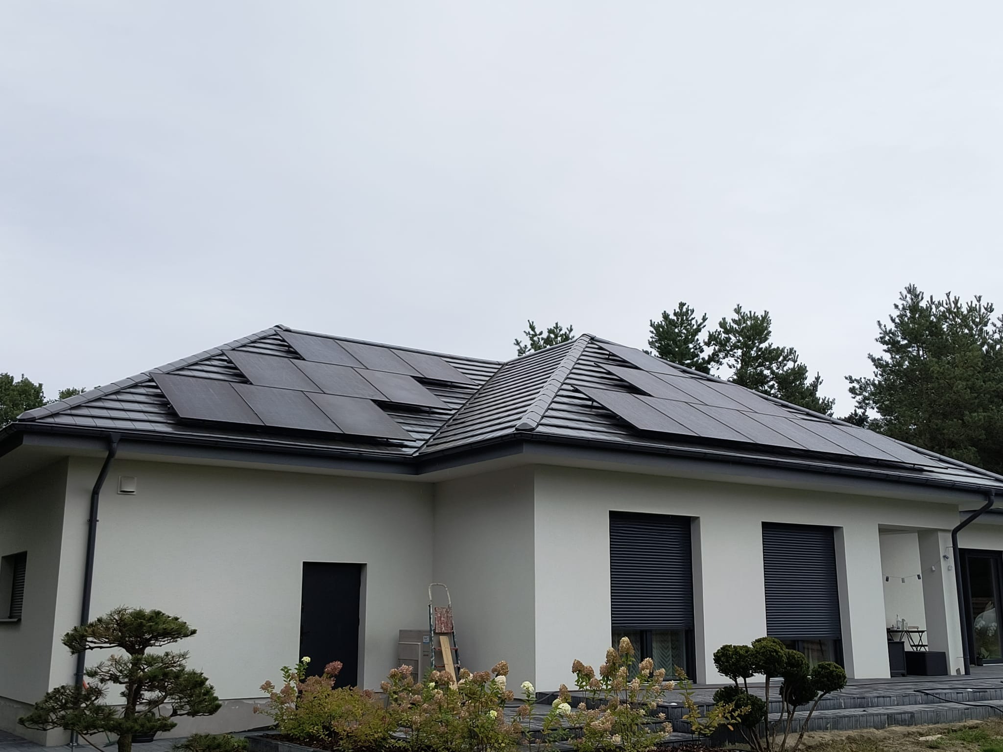 Dom jednorodzinny z szarym dachem i panelami słonecznymi. Widoczne rolety zewnętrzne i ozdobne krzewy w ogrodzie.