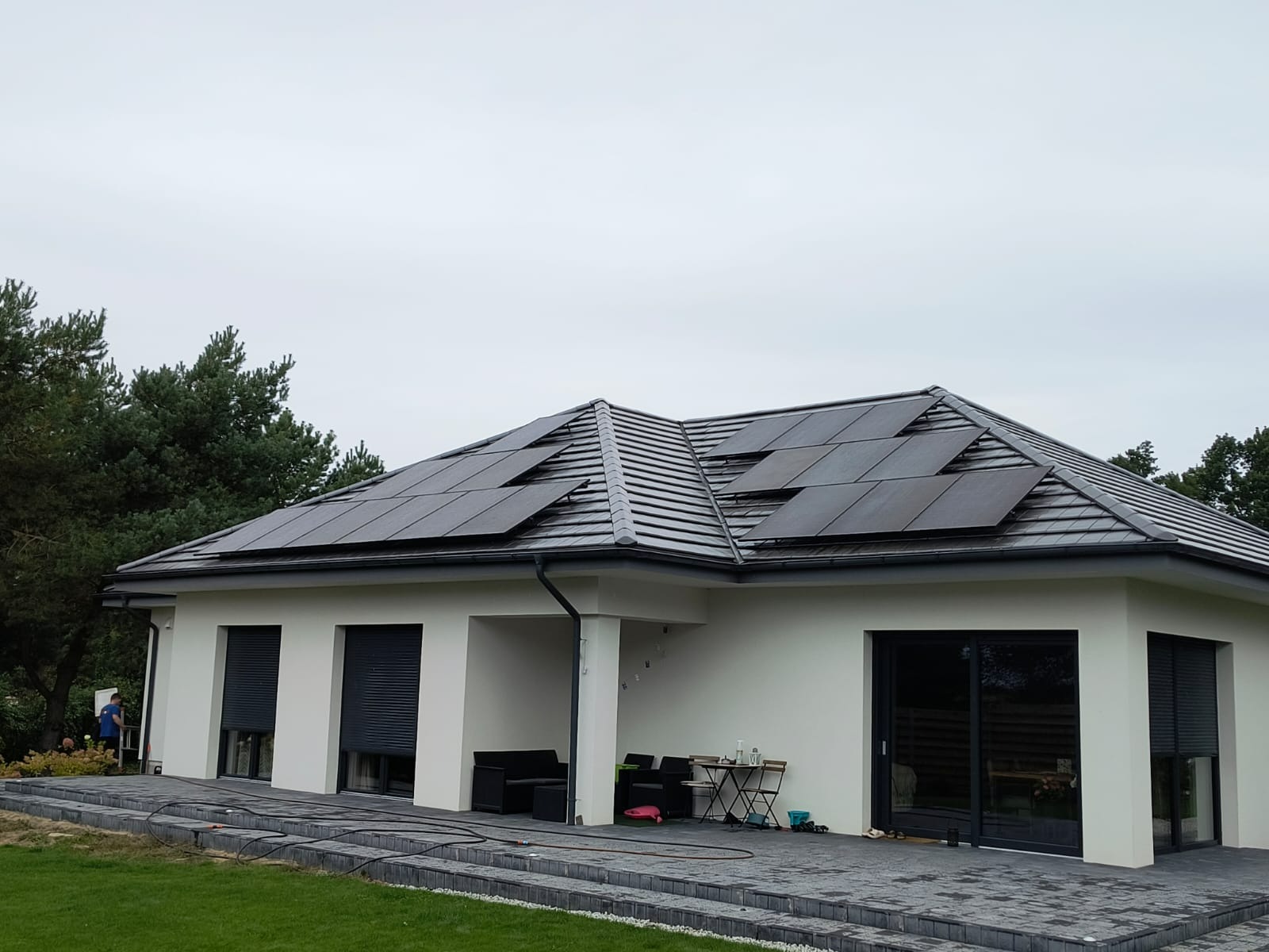 Nowoczesny dom jednorodzinny z panelami fotowoltaicznymi na dachu. Widoczne czarne panele na szarej dachówce. Biała elewacja budynku i szare elementy wykończeniowe.