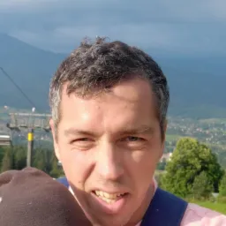 Autoportret mężczyzny z szarymi włosami na tle górskiego krajobrazu z wyciągiem narciarskim i miasteczkiem w dolinie, pochmurne niebo.