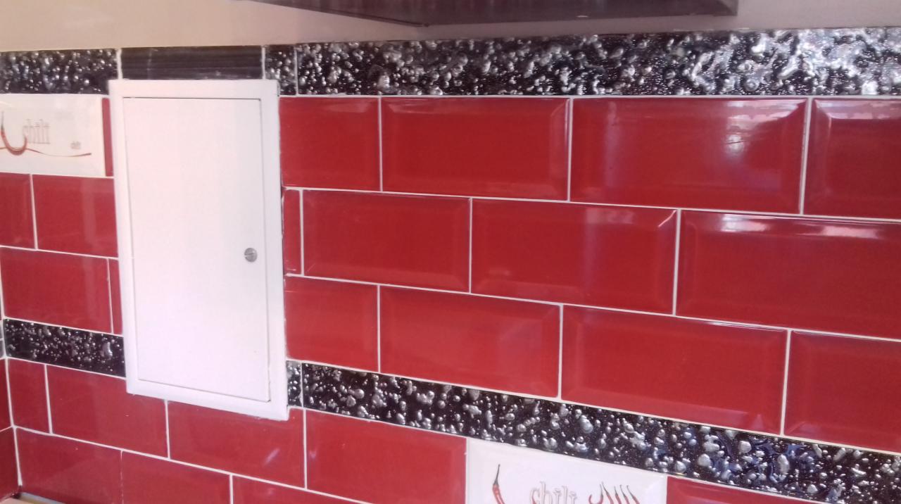 Czerwone, błyszczące płytki ułożone w rzędach, przedzielone ozdobnym pasem z motywem srebrnych kropek na czarnym tle, z białymi płytkami dekoracyjnymi po bokach i białymi drzwiczkami rewizyjnymi.