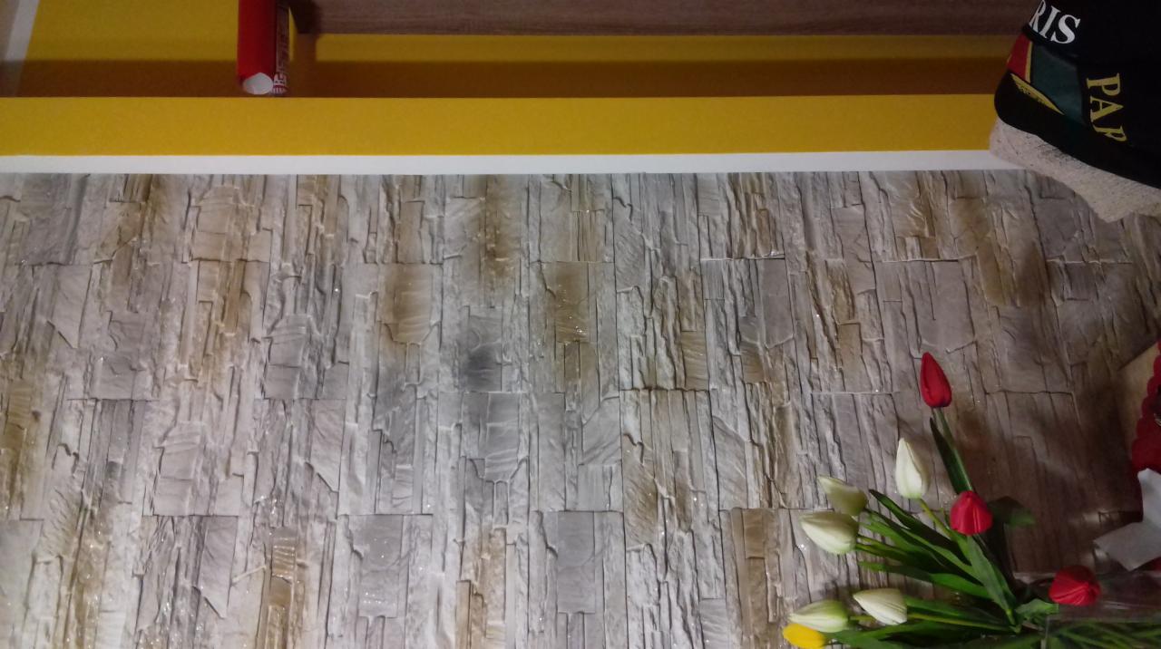 Ściana wykończona dekoracyjną okładziną imitującą kamień w odcieniach szarości i beżu, widoczne sztuczne tulipany w wazonie oraz fragment półki z żółtym obramowaniem.