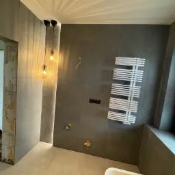 Szara łazienka w trakcie wykończenia: płytki na ścianach, grzejnik drabinkowy, wiszące lampy, widoczne instalacje, surowy otwór drzwiowy, fragment wanny.