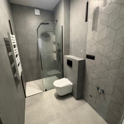 KRIS Nowe Wnętrza remont z pasją - Nowoczesna łazienka w odcieniach szarości z kabiną prysznicową typu walk-in, wiszącą toaletą i grzejnikiem drabinkowym. Ściana z geometrycznym wzorem płytek.