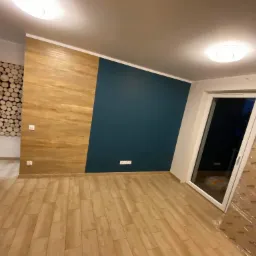 Pokój po remoncie: ściana z drewnianą okładziną i pomalowaną na granatowo, podłoga wyłożona panelami imitującymi drewno, drzwi balkonowe w folii ochronnej.