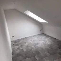Poddasze po remoncie: szare płytki podłogowe imitujące kamień, białe ściany, okno dachowe wpuszczające naturalne światło, widoczne gniazdka elektryczne.