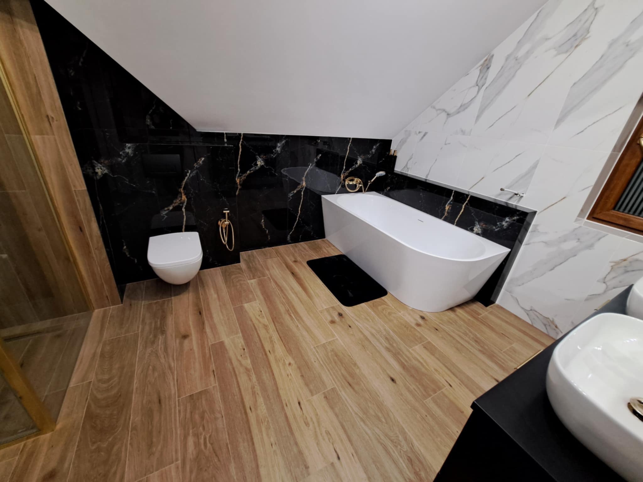 Nowoczesna łazienka na poddaszu z drewnopodobną podłogą, czarnymi i białymi marmurowymi płytkami, wolnostojącą białą wanną oraz umywalką nablatową.