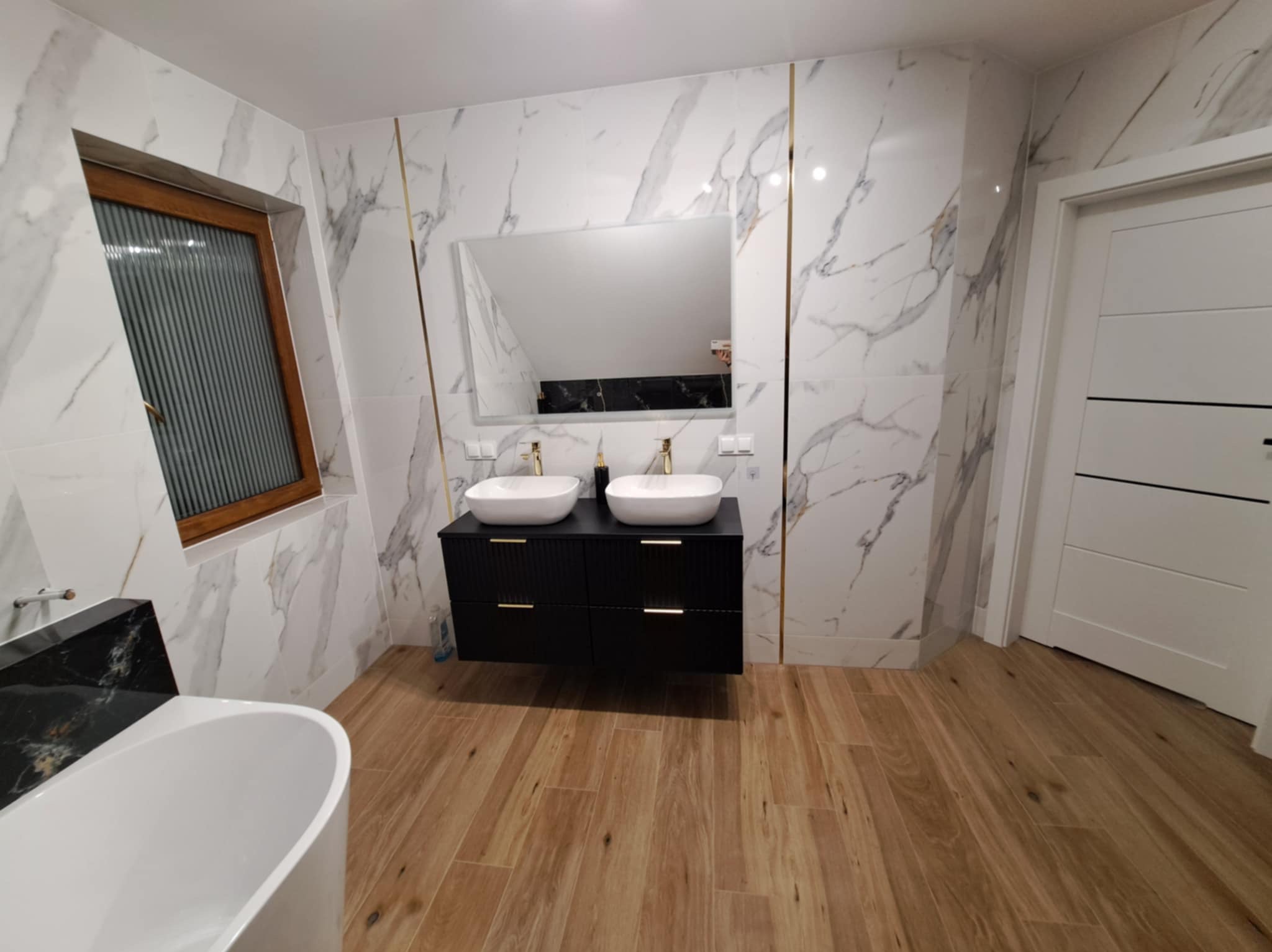 Nowoczesna łazienka z marmurowymi płytkami, drewnopodobną podłogą, czarną szafką z dwiema umywalkami i złotymi akcentami na płytkach oraz oknem z drewnianą ramą.