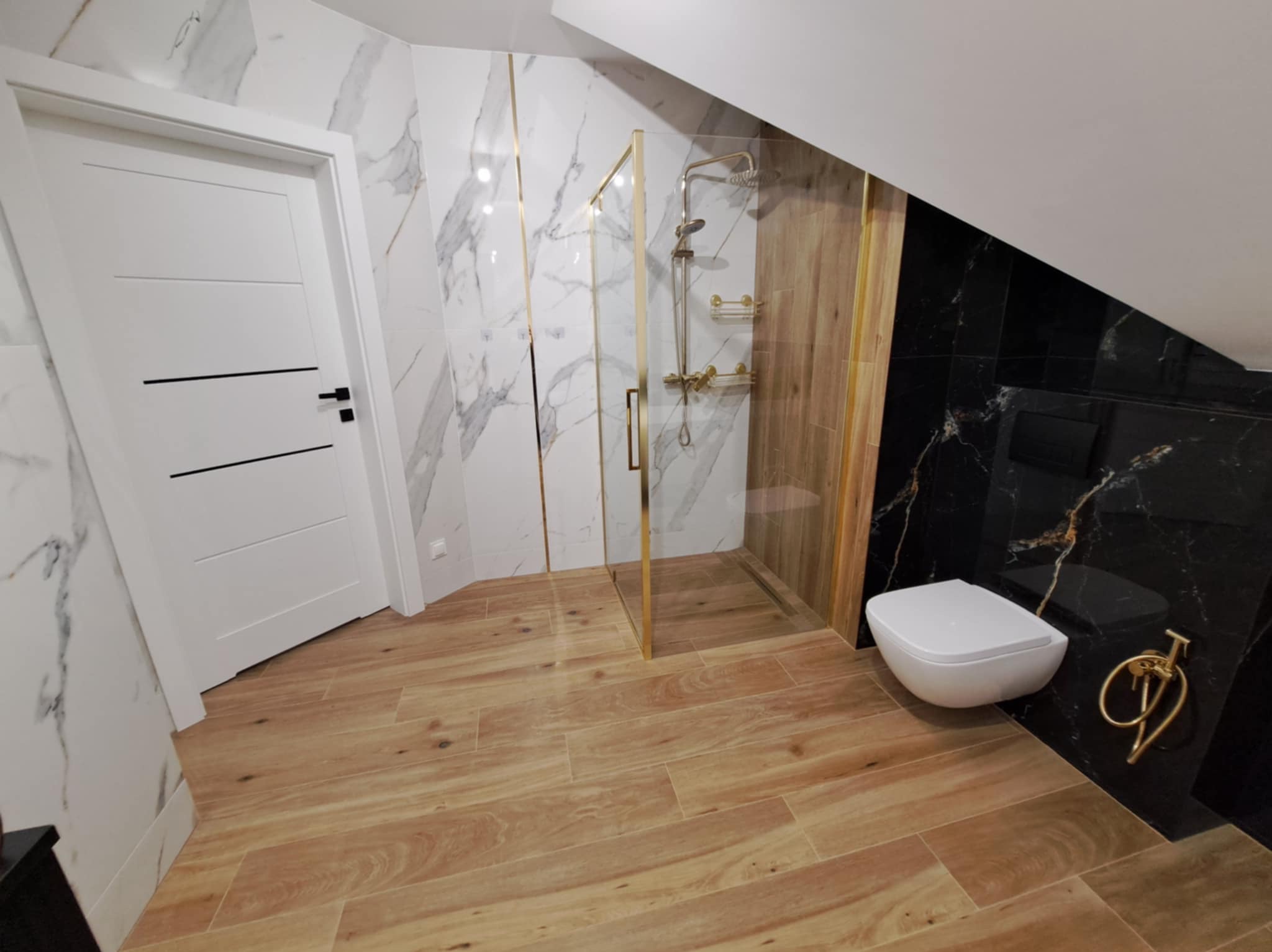 Nowoczesna łazienka na poddaszu z kabiną prysznicową ze złotymi detalami, wiszącą toaletą i marmurowymi płytkami.
