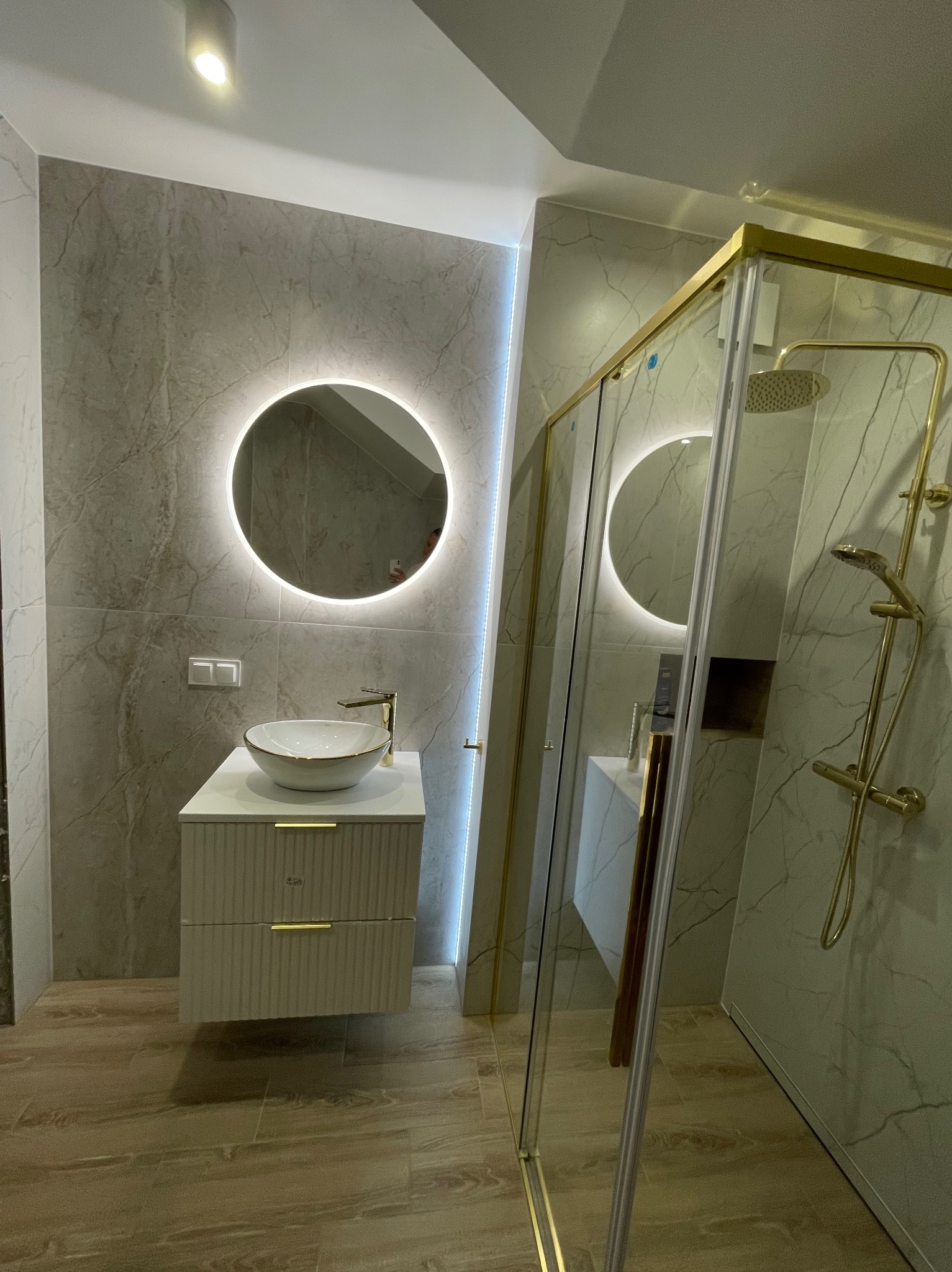 Nowoczesna łazienka z marmurowymi płytkami, okrągłym lustrem z podświetleniem LED, białą umywalką nablatową z złotą baterią i kabiną prysznicową ze złotymi profilami.