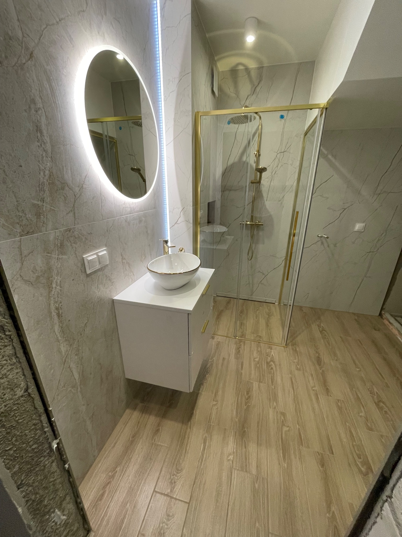 Nowoczesna łazienka z owalnym lustrem LED, kabiną prysznicową ze złotymi ramami i umywalką nablatową na białej szafce. Płytki imitujące marmur na ścianach i podłodze.