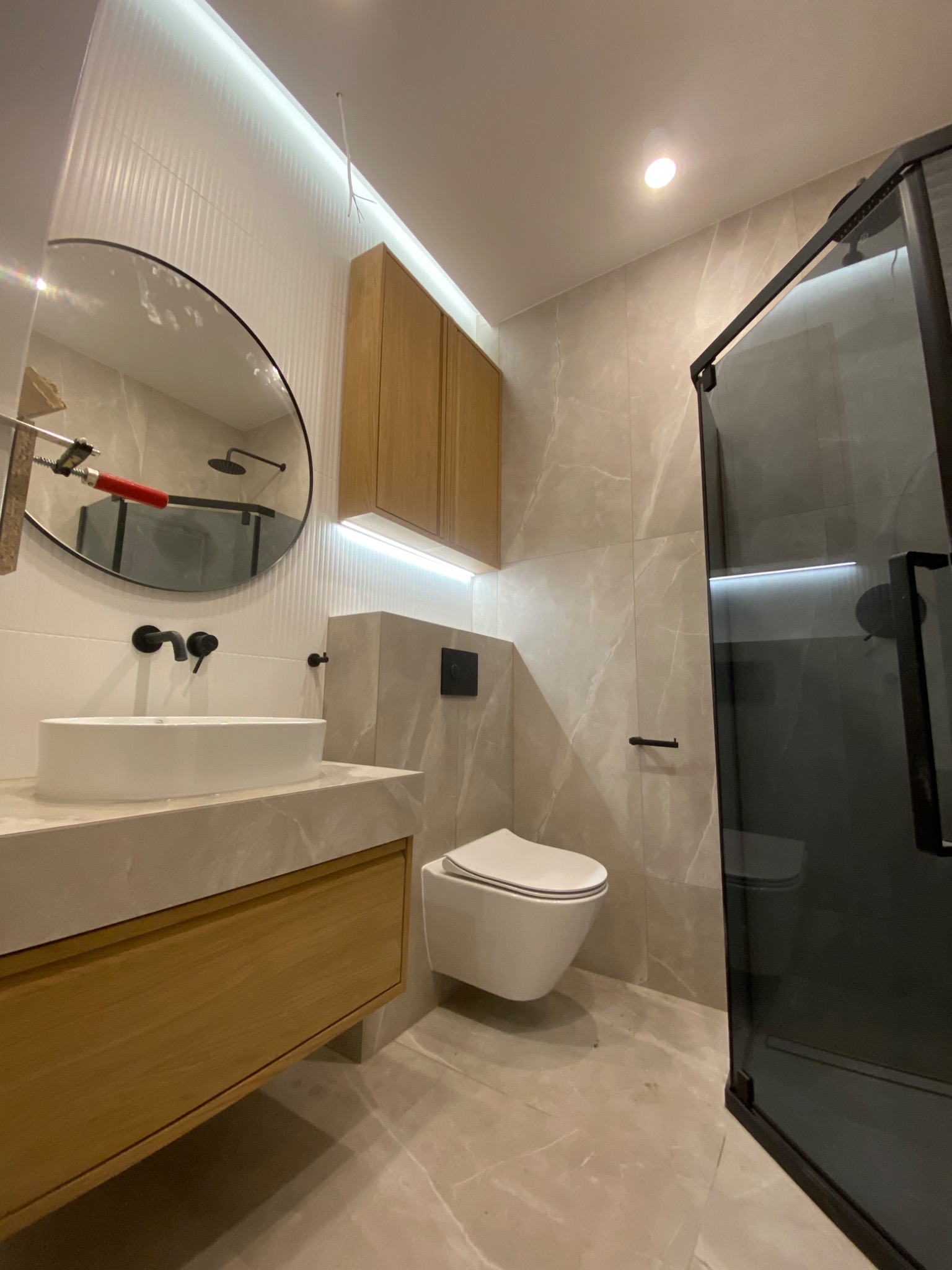 Nowoczesna łazienka z okrągłym lustrem, drewnianą szafką pod umywalką, podwieszaną toaletą i kabiną prysznicową z czarnymi ramami, oświetlona ledami.