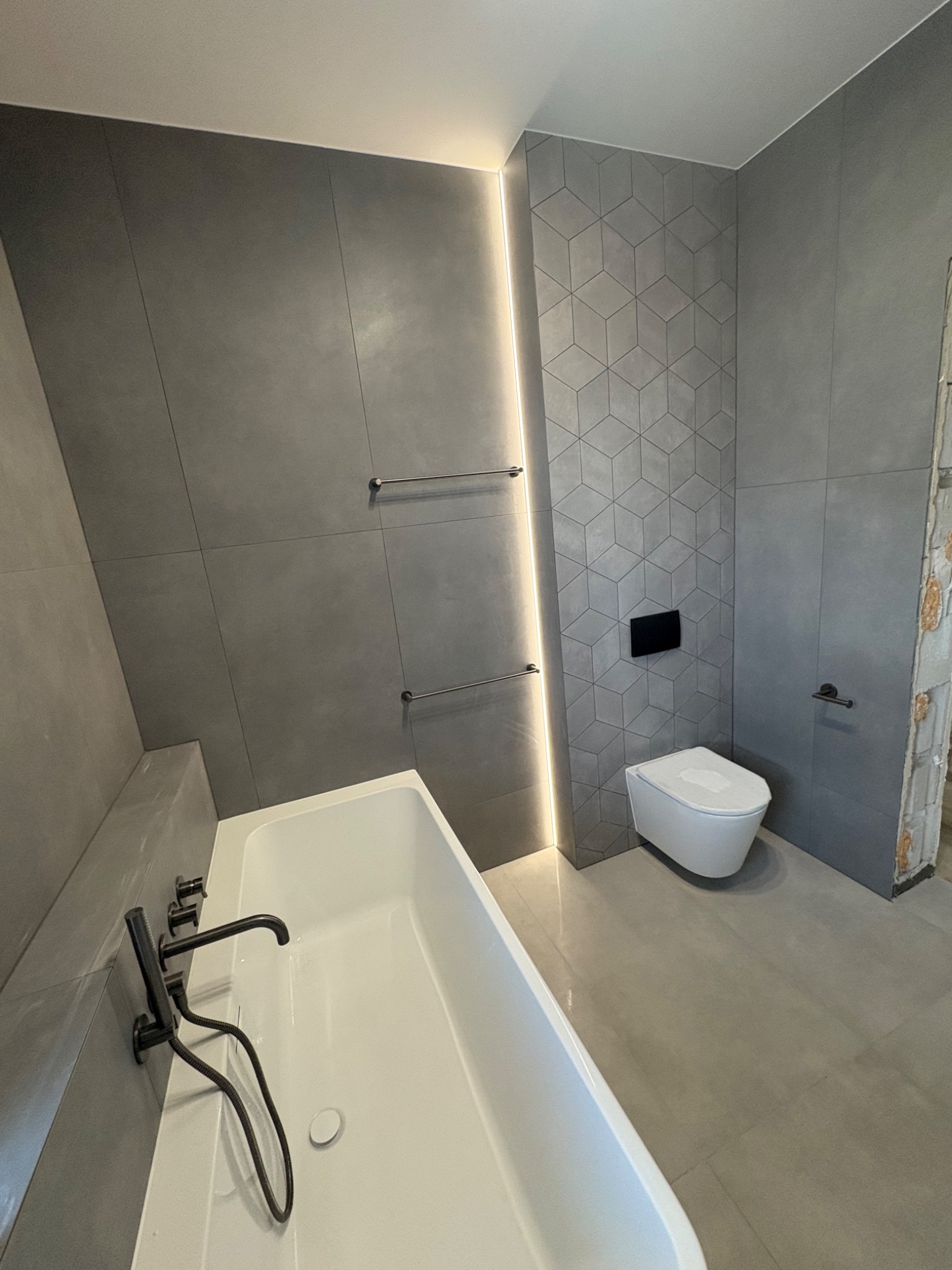 Nowoczesna łazienka w szarościach: geometryczne płytki, podświetlenie LED, minimalistyczna armatura i wisząca toaleta. Częściowo widoczna wanna.