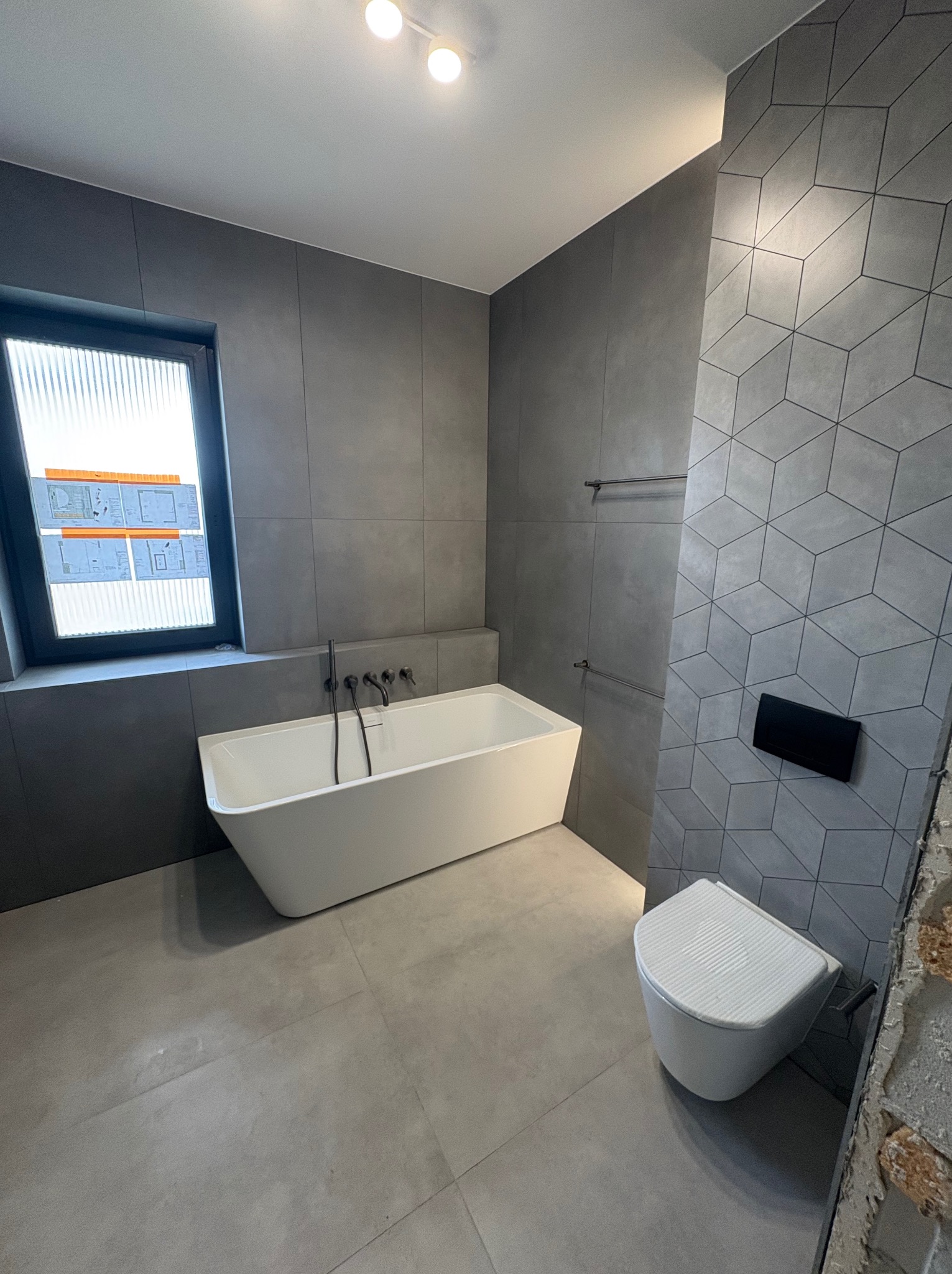 Nowoczesna łazienka w szarościach: geometryczne płytki, wolnostojąca wanna z czarną armaturą, podwieszana toaleta i okno z widocznymi planami architektonicznymi.