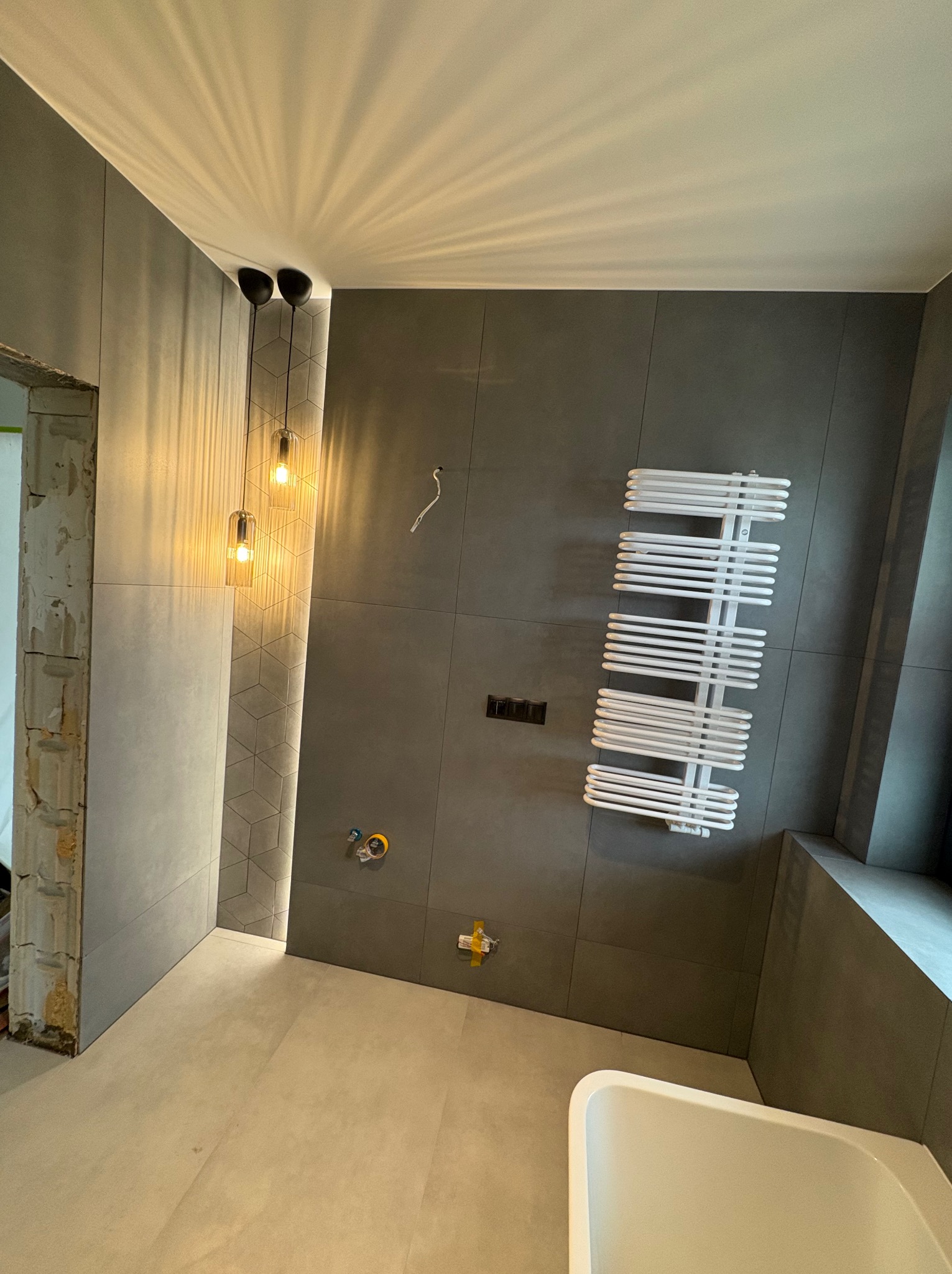 Szara łazienka w trakcie wykończenia: płytki na ścianach, grzejnik drabinkowy, wiszące lampy, widoczne instalacje, surowy otwór drzwiowy, fragment wanny.