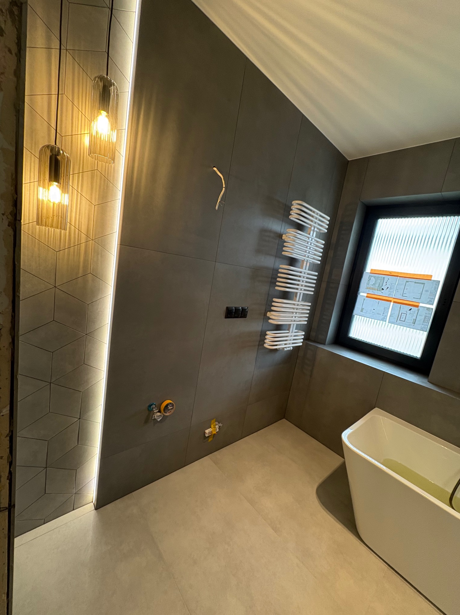 Nowoczesna łazienka z szarymi płytkami, dekoracyjnym oświetleniem LED, grzejnikiem drabinkowym i białą wanną. Widoczne przyłącza wodne.