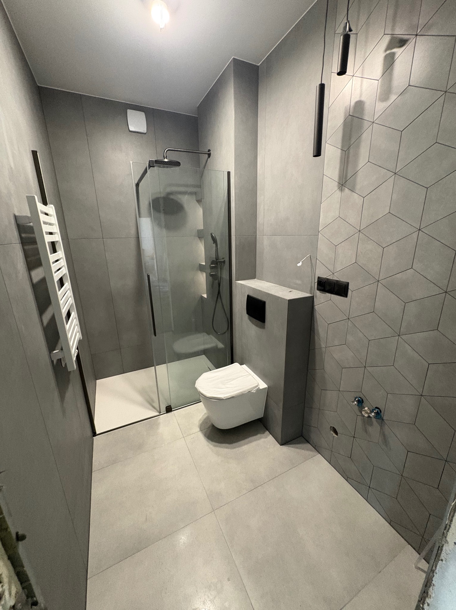 Nowoczesna łazienka w odcieniach szarości z kabiną prysznicową typu walk-in, wiszącą toaletą i grzejnikiem drabinkowym. Ściana z geometrycznym wzorem płytek.