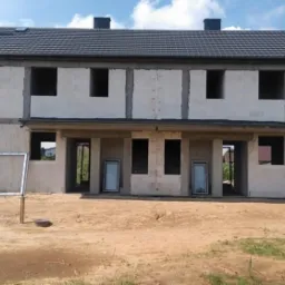 Surowy stan dwukondygnacyjnego budynku z pustaków z widocznymi otworami okiennymi i drzwiowymi, fragment białego busa po lewej stronie, okno w ramie na pierwszym planie, ziemisty teren przed...