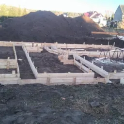 Przygotowanie szalunków pod fundamenty domu jednorodzinnego, widoczne drewniane formy na ziemi, w tle sterta ziemi i zabudowania gospodarcze.