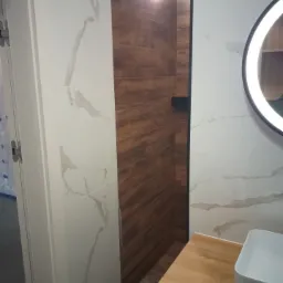 Nowoczesna łazienka z kabiną prysznicową wyłożoną płytkami imitującymi drewno, kontrastująca z białymi płytkami z marmurowym wzorem na pozostałych ścianach, okrągłe lustro z podświetleniem LED...