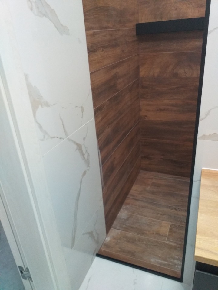 Wykończona wnęka prysznicowa z brązowymi płytkami imitującymi drewno, kontrastująca z białymi płytkami imitującymi marmur na sąsiedniej ścianie, czarna listwa wykończeniowa.