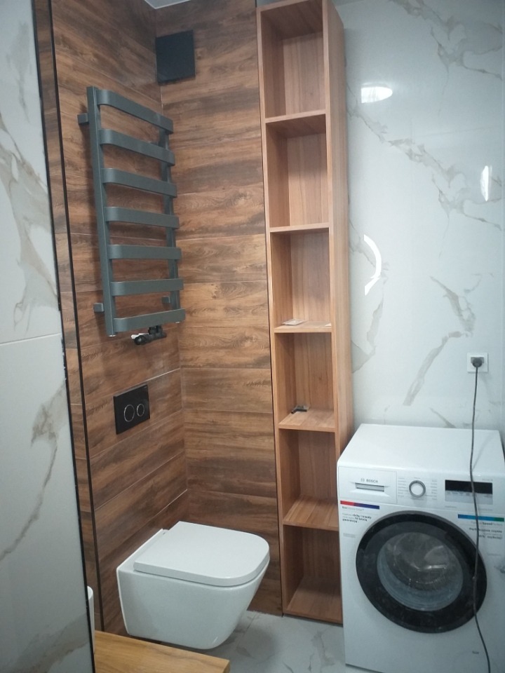 Nowoczesna łazienka po remoncie: wisząca toaleta, grzejnik drabinkowy, drewniany regał i pralka Bosch na tle płytek imitujących drewno i marmur.